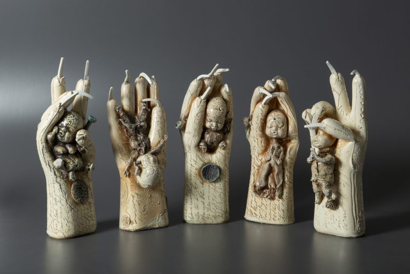 five sculptures of hands with pointy nails holding tiny babies in their palms.