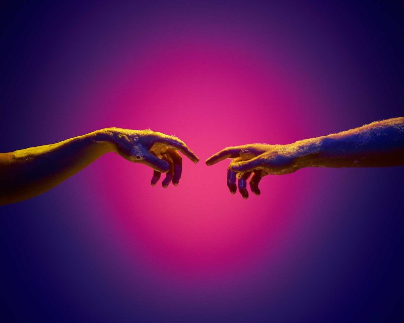 Two soapy hands reaching their forefingers towards each other in front of a radially lit pink and purple background