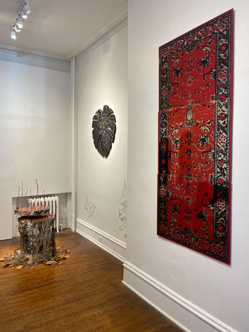 Photo shows a corner of a room in a gallery with a rug on a wall, a small object in the background and track lighting on the ceiling.