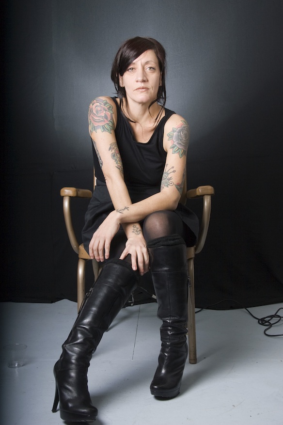 photo of woman sitting in chair, black hair, black dress, black boots, arms crossed at wrists, looking concerned