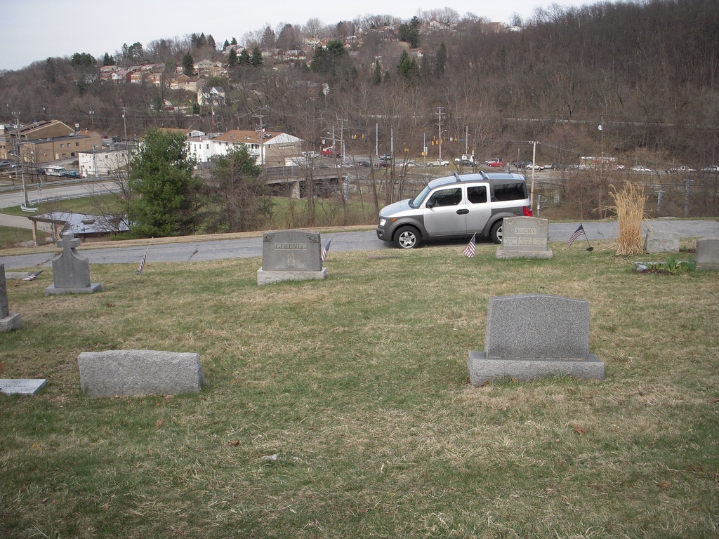 Graveyard with road, grey car, tree-topped hill in background. 