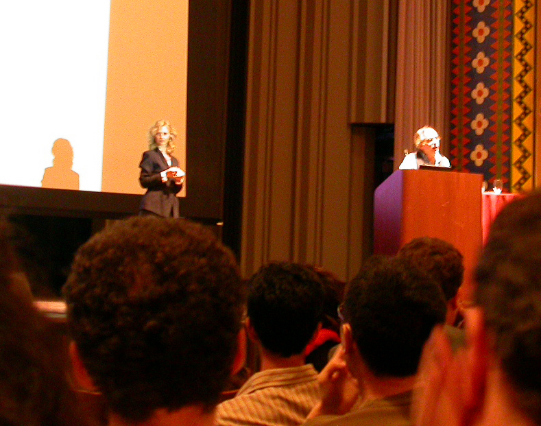 A man stands at a podium giving a lecture and a woman stands off to the side in the background