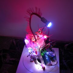 Huang, Shih Chieh's tiny robot on a turntable. It flashes and tweets and is friendly as a video arcade game. Materials include a toy gun and what look like Tupperware containers, in addition to plastic handcuffs, lights and circuitry.