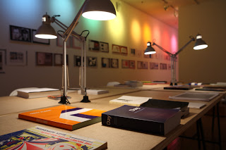 Books laid out in a reading room, complete with individual reading lights. Double Spread stretches across the back wall.