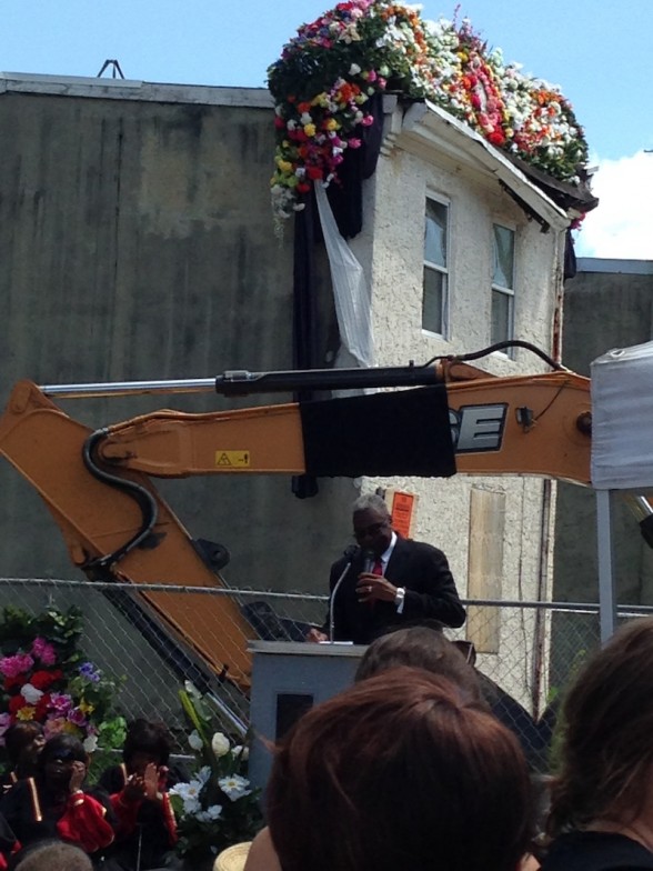 Pastor Harry Moore Sr. of Mount Olive Baptist Church, giving the eulogy for 3711 Melon St. Notice black "arm band" on the excavator