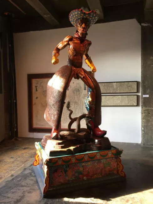 A sculpture of a cowboy with an erection in chaps and a big cowboy hat. He has clenched fists and his legs are bowed. He stands upon a painted plinth. He is rendered in wood.