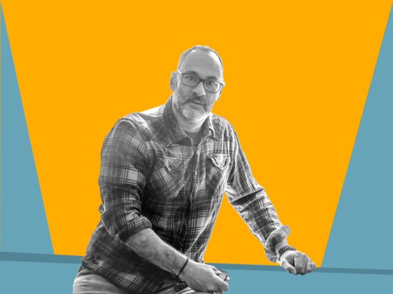 Portrait of a man with a beard and glasses leaning on a table you don't see with a bright orange and teal background.