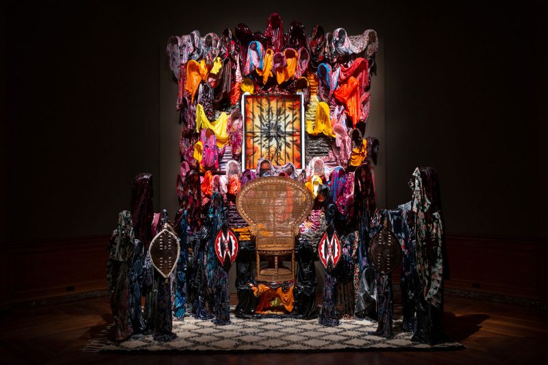 Kevin Beasley, "Chair of the Ministers of Defense," 2016, Polyurethane resin, wood, acoustic foam, jeans, trousers, du-rags, altered t-shirts, altered hoodies, guinea fowl feathers, wrought iron window gate, vintage Beni Ourain Moroccan rug, kaftans, housedresses, Maasai war shields, Zulu war shields, and vintage peacock rattan chair. Overall: 176 1/4 × 162 × 84 in. The Joyner/Giuffrida Collection, San Francisco and Rennie Collection, Vancouver. Mitro Hood Photography