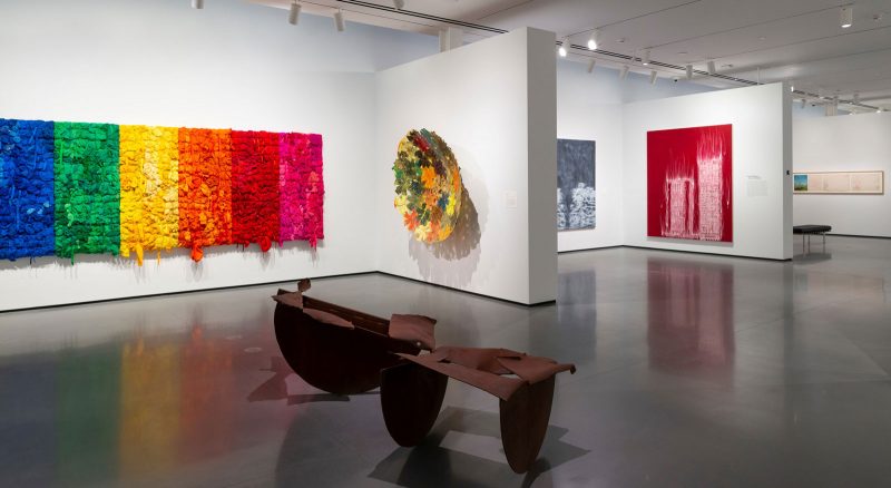 Installation view, "Generations: A History of Black Abstract Art" at The Baltimore Museum of Art. Pictured: Shinique Smith, "Black, Blue, Green, Yellow, Orange, Red, Pink," 2015, Clothing, fabric, and ribbon on wood panel, 72 × 252 × 6 in., The Joyner / Giuffrida Collection. Kevin Beasley, "Bronx Fitted," 2015, New Era® Yankee fitted hats, bandanas, polyurethane resin, and television mount 70 × 70 × 16 in., The Joyner / Giuffrida Collection. Gary Simmons, " In To You Like A Train," 2000, Oil on canvas, 84 × 84 in., The Joyner / Giuffrida Collection. Gary Simmons, "Double Cinder," 2007, Pigment, oil paint, and cold wax on canvas 102 1/2 × 84 1/4 in. The Joyner / Giuffrida Collection. Photo courtesy: Mitro Hood.