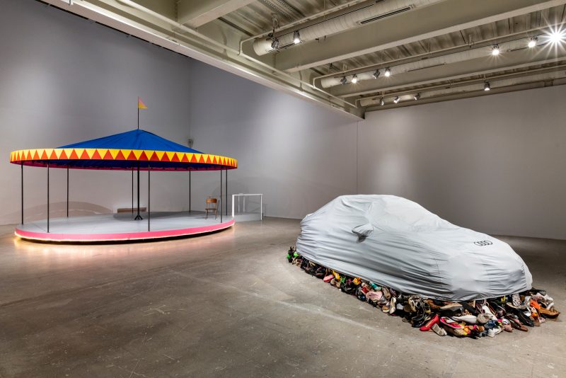 In the background and the left, a colorful carousell with a chair on it that rotates. In the foreground and right, a pile of shoes that are hidden under a car cover, taking the shape of a car. Shoes spill out in all of the sides.