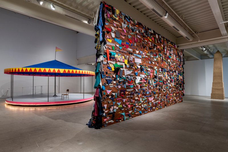 Background: in the left, a carousel with a chair sitting on it. On the right, an obelisk that reaches to the ceiling of the institute of contemporary art. Foreground, between both sculptures: A wall made of bricks with bits of clothing sticking out in between each of the bricks.