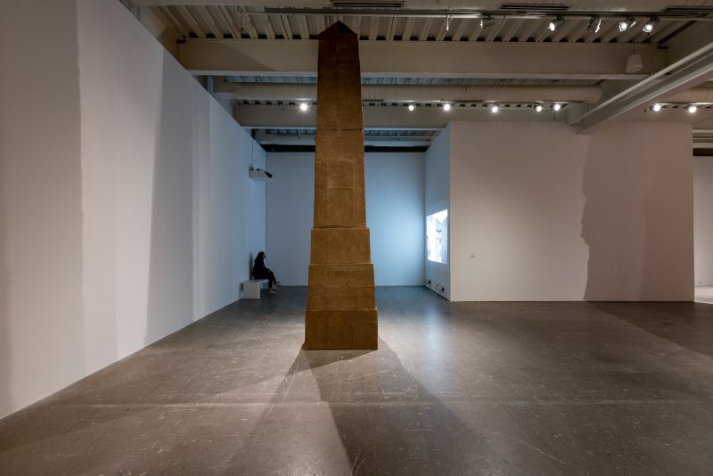 A big obelisk that reaches the top of the ceiling. Behind it, a person watches a film about the making of the obelisk and the history of the obelisk, which is projected on a wall opposite to them.