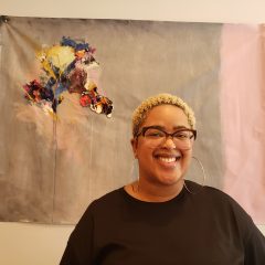 A'Driane Nieves in a black shirt, wearing glasses and hoop earrings, posing in front of a gray, abstract painting with an explosively colorful focal point, painted on paper.
