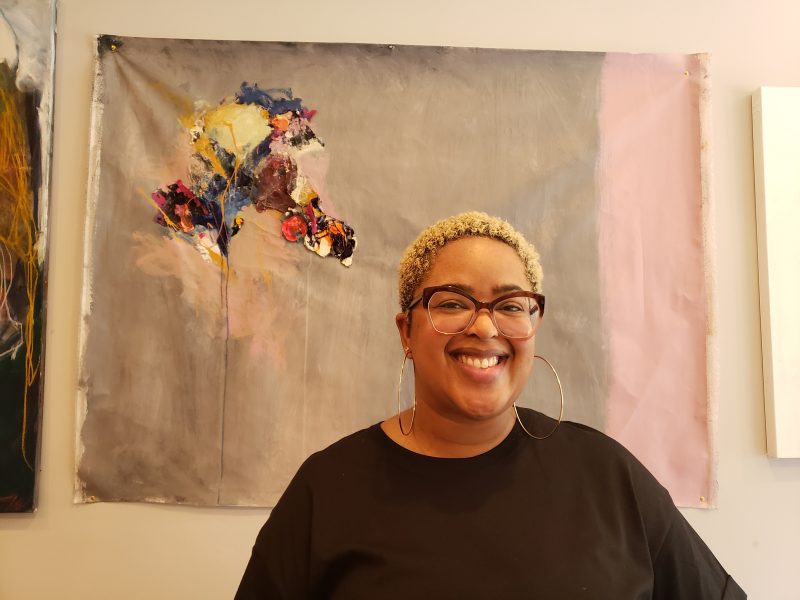 A'Driane Nieves in a black shirt, wearing glasses and hoop earrings, posing in front of a gray, abstract painting with an explosively colorful focal point, painted on paper.