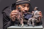 Artist Acori Honzo holds and positions two dolls sculpted by himself of two men speaking to one another on a park bench also sculpted by the artist