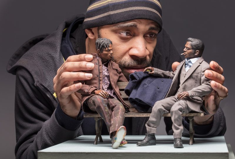 Artist Acori Honzo holds and positions two dolls sculpted by himself of two men speaking to one another on a park bench also sculpted by the artist.
