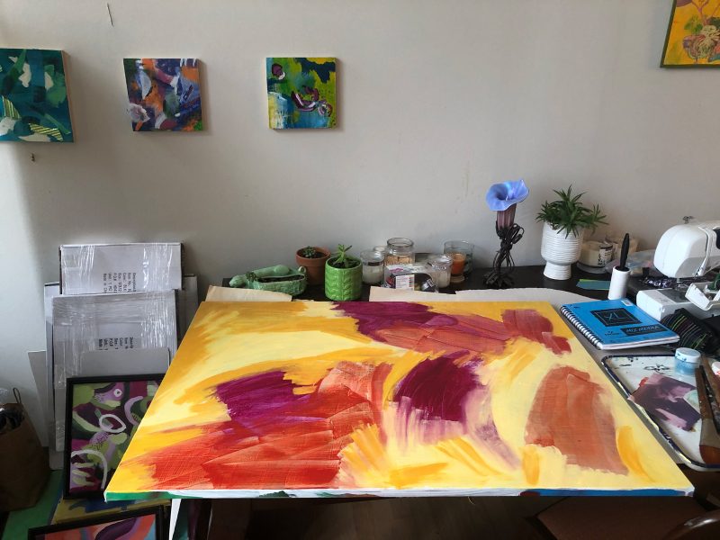 Artist studio with a large painting in progress on a table with plants and supplies. There are two small paintings on the wall.