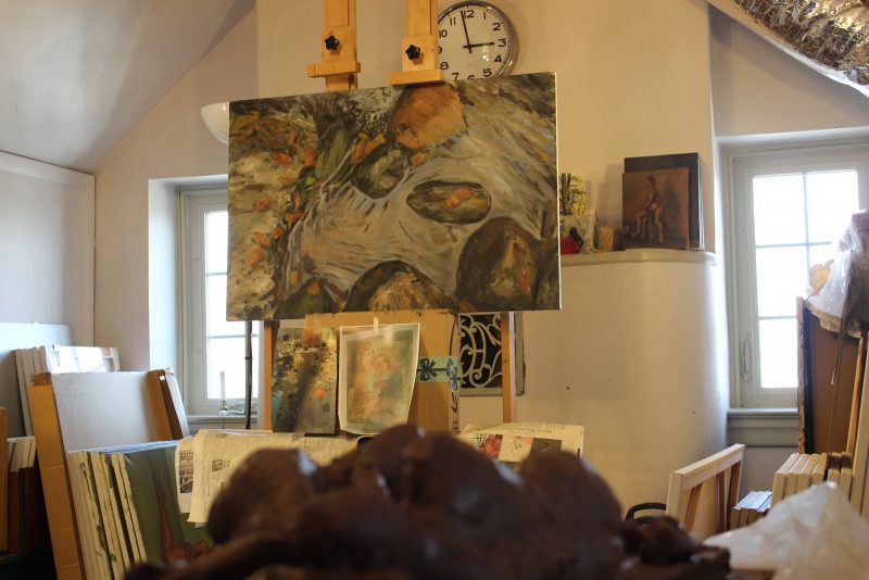 Painting of rocks on an easel in a home studio containing lots of painting canvases stacked up on the floor and paintings hangin on the walls. 