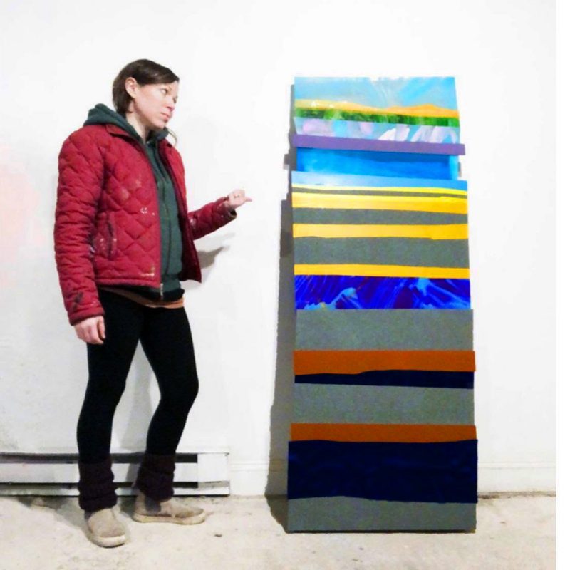 Rita Leduc stands next to their large work of art. The art is tall and contains layers of colors in a stripe pattern, some of which contain additional decorate marks.