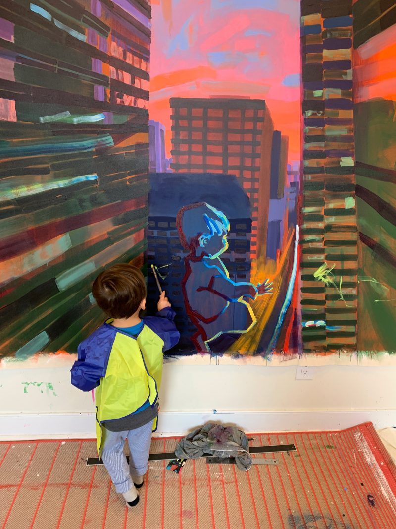 Painting of a colorful cityscape and an outline of a young boy hanging up on the wall. A young boy looking at the painting.
