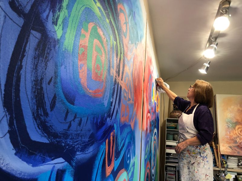 Jacqueline Unanue in her studio, painting two large scale abstract paintings that are blue, red, green, and orange.