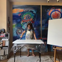 Jacqueline Unanue in her studio, preparing surfaces to paint on, standing in front of her large, blue and green and red abstract paintings.