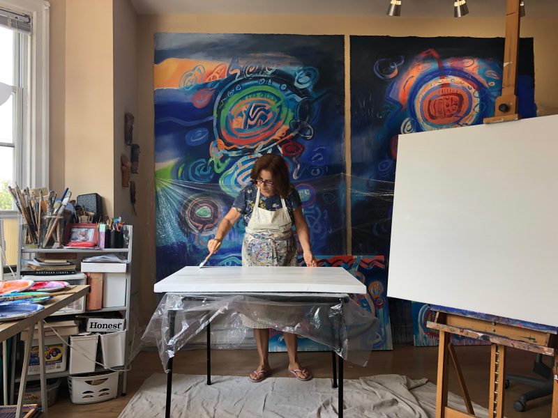 Jacqueline Unanue in her studio, preparing surfaces to paint on, standing in front of her large, blue and green and red abstract paintings.