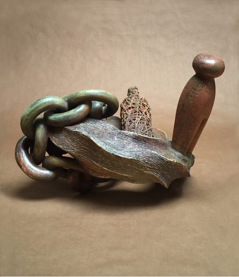 Wooden carved sculpture of a metal chain around a wooden platform with a clothes pin-like shape sticking out of the right hand side. 