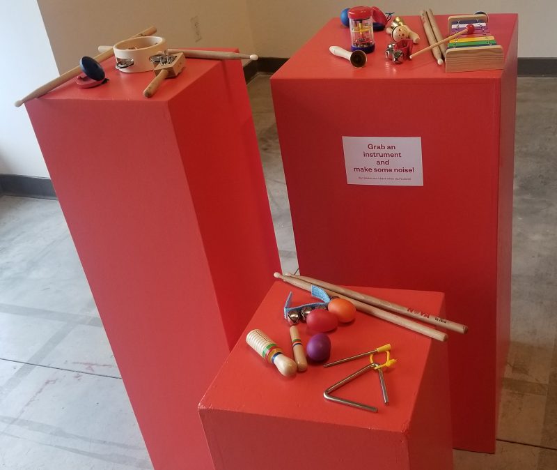 Red pedestals with triangles, shakers, tambourines, and drumsticks, with a note that says "Grab an instrument and make some noise!"