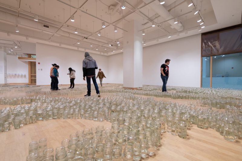 People walking through a gallery near glass jars containing water arranged in a pattern on a gallery floor.