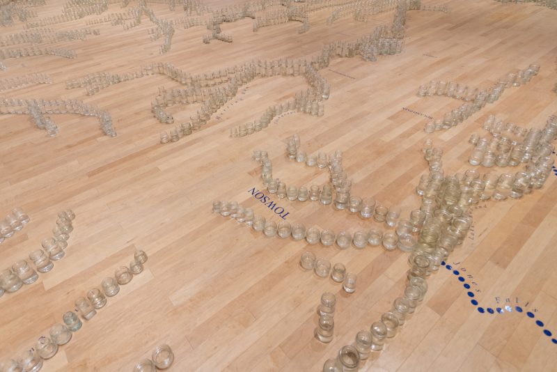 Glass jars containing water arranged in a pattern with text: "Towson" and other vinyl patterns on a gallery floor.