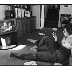 Young adult Judy Gelles sitting on the floor watching TV on a small black and white TV in a living room.