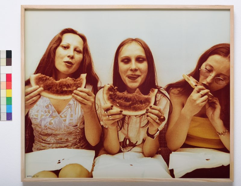 Three young women eating watermelon in a line, all wearing white skirts.