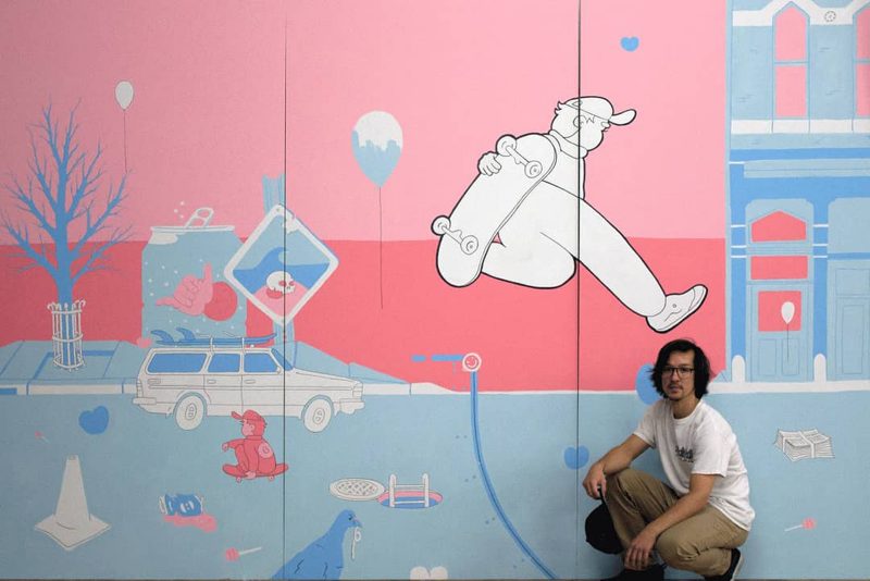 James Bonney kneeling in front of their pink and blue painting of a city scape with a skateboarder in the air.