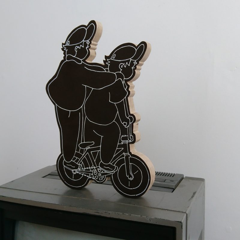 Wooden sculpture of a young person riding a bike with someone else riding on their pegs
