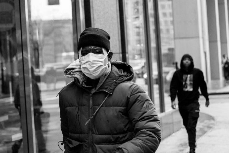 Black and white photograph of a man walking down the street with a mask over his mouth.