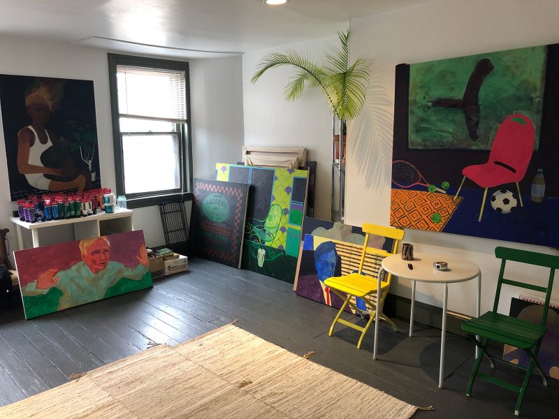 Travis's art studio, filled with plants, paintings on the walls, and artwork in stacks on the floor leaning against the walll.