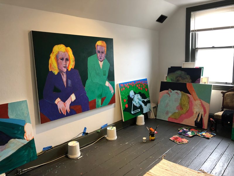 Travis's studio, with a large painting of two figures in suits hanging on the wall and stacks of paintings in a corner.