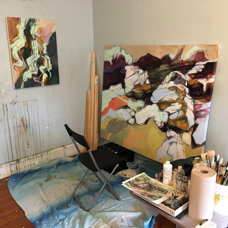 Painting of abstracted rock formations leaning against the wall in a studio with a chair in front and a table of supplies to the right of it.