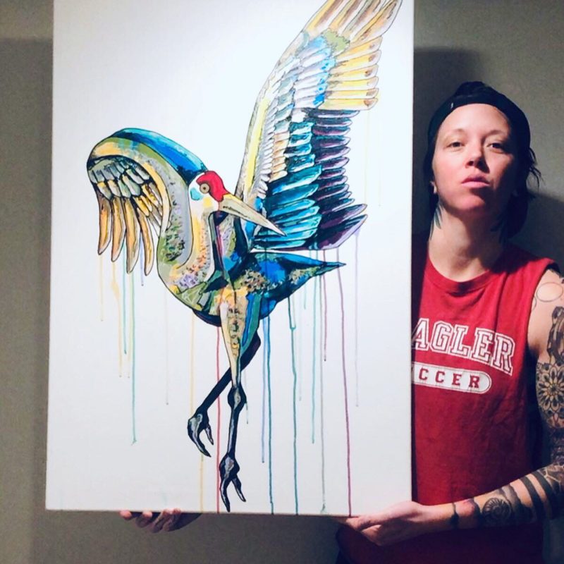 Tiff Urquhart posing with a large, colorful mixed media drawing of a sandhill crane on a white background.