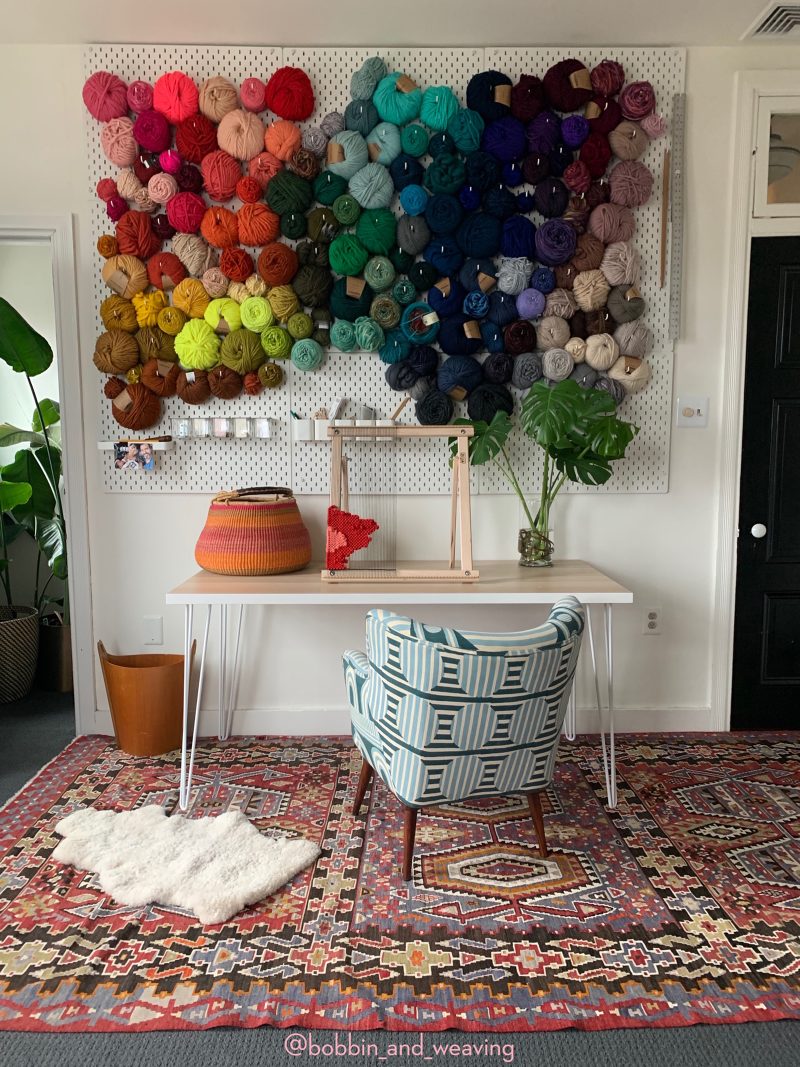 A pegboard full of yarn on the wall of an at home studio with a deck, a loom and a chair. 