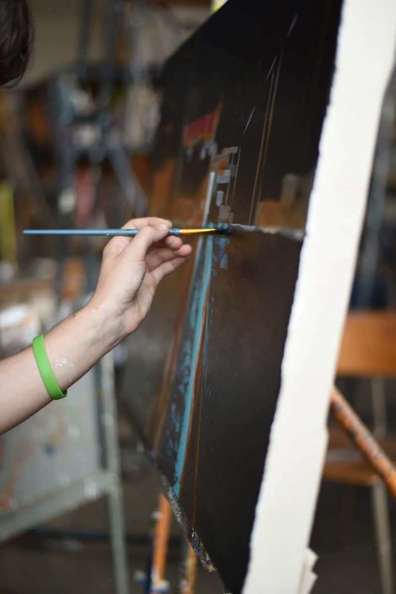 Taylor Bielecki's hand from the side, holding a paintbrush and painting on a large rectangular canvas.