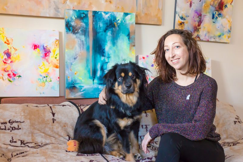 Juli sitting on a couch in front of their paintings, posing with their dog.