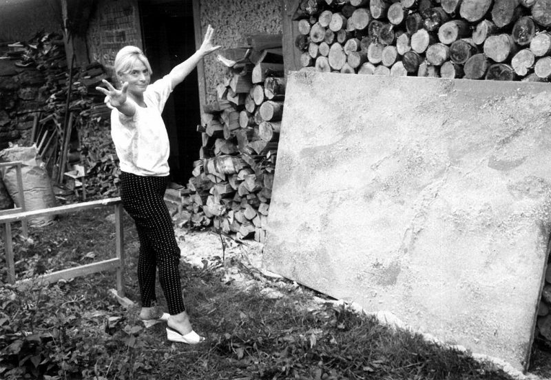 Natalie posing with a peace sign in front of a large canvas propped against a stack of logs.