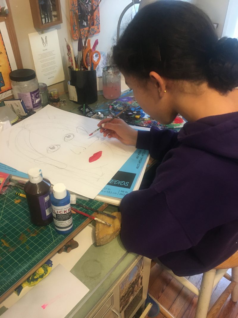 Young child sitting at a desk drawing a face with red lips.