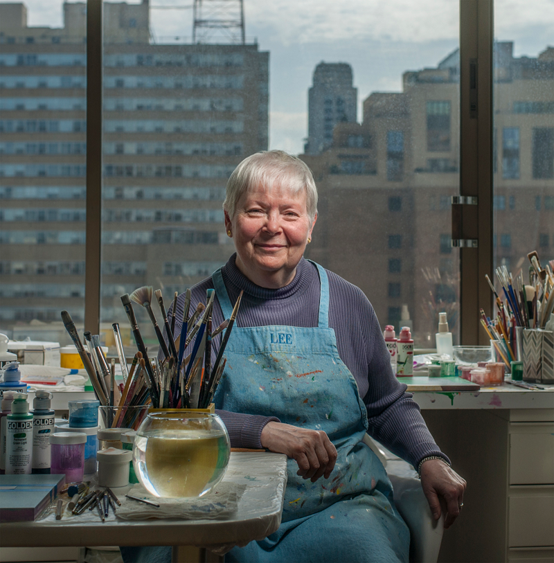 Linda Lee Alter sitting in their studio holding a brush in their hand, wearing an apron and sitting at their desk.