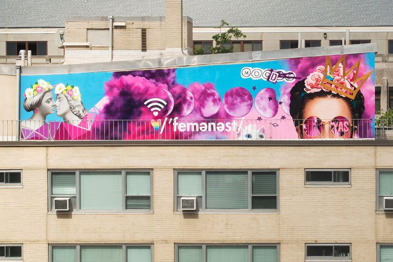 Large billboard or mural style painting of a woman, purple clouds, and text "femenest" on the top of a city building