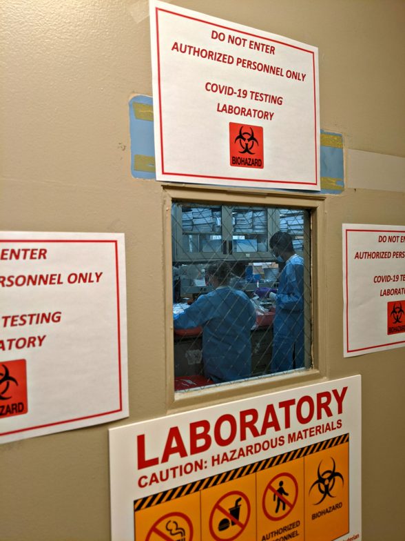 Two people working in a lab photographed from outside the door with Hazardous labels on the door.
