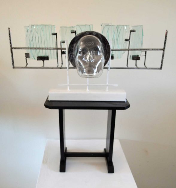 Sculpture of a mask with metal rods coming out holding three glass squares. 