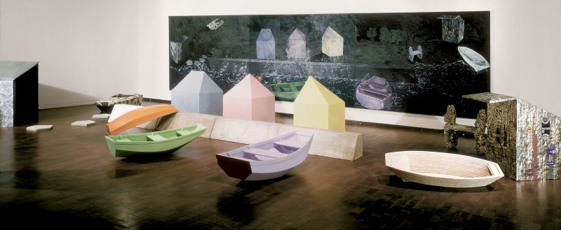 Sculptures of pastel colored wooden boats in front of a photo of water